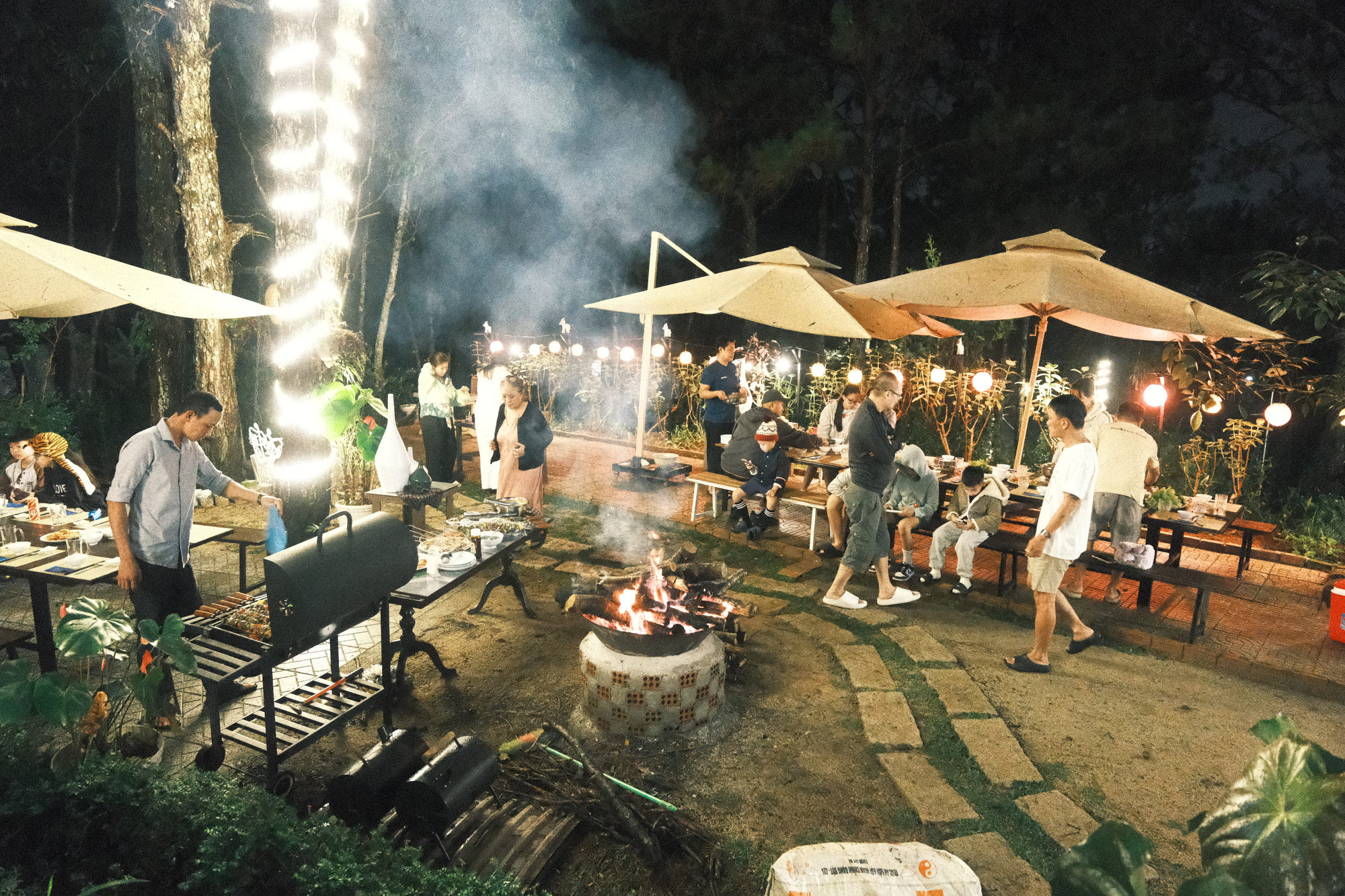 Tiệc BBQ gala của đoàn dưới tán thông với bếp nướng, dù lớn và dây đèn tại GIA AN